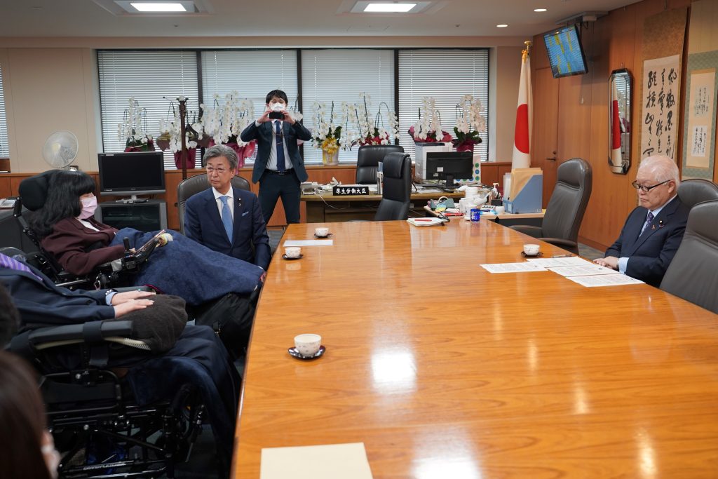 画面左側に、発言する木村議員。その右隣に石井委員長が着席。木村議員と机をはさんで正面に座る武見大臣が、要望書に目を落としながら木村議員の発言を聞いている。