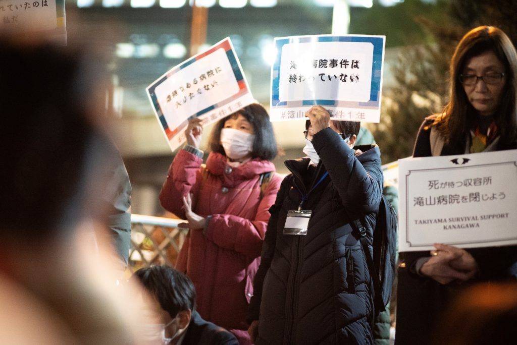 夜間の都庁前でプラカードを掲げて立つ女性3人が並ぶ。プラカードには「虐待のある病院に入りたくない」「滝山病院事件は終わっていない」「死が待つ収容所滝山病院を閉じよう」と書かれている。